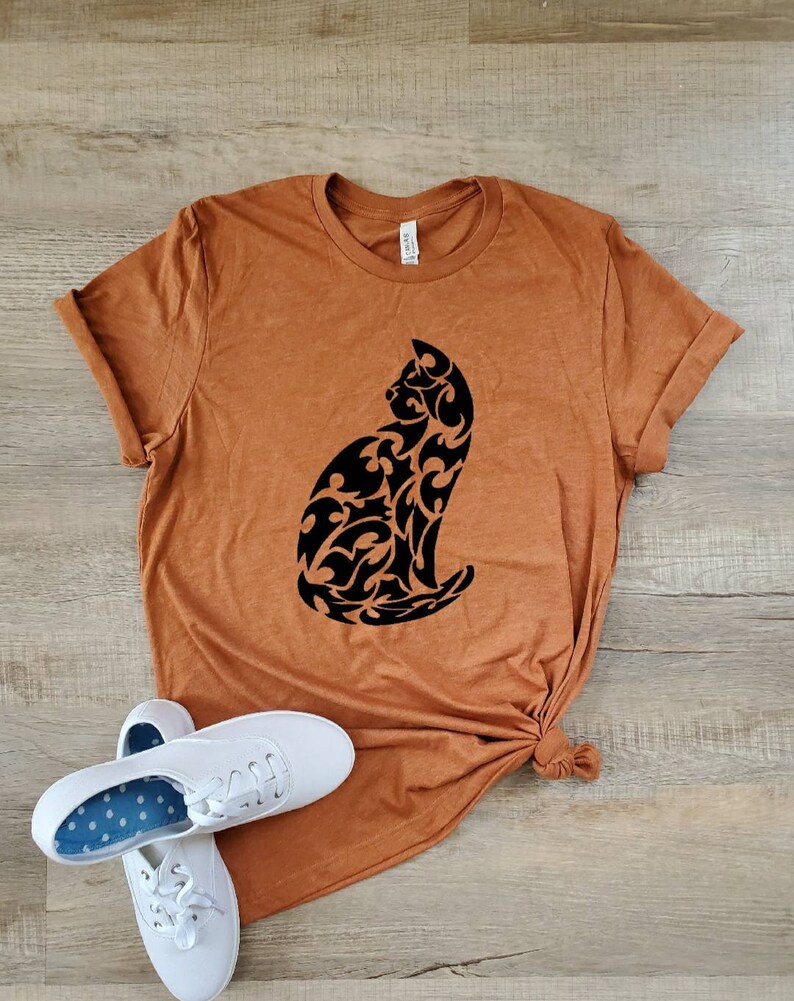 May include: A rust-colored t-shirt with a black tribal cat design. The shirt is made of a soft, comfortable material and has a relaxed fit.
