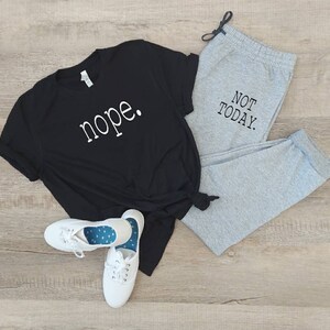 Puede incluir: Una camiseta negra con la palabra "nope" impresa en blanco en la parte delantera, combinada con pantalones de ch&aacute;ndal grises con el texto "NOT TODAY" impreso en negro en la pierna. El conjunto est&aacute; colocado sobre un suelo de madera.