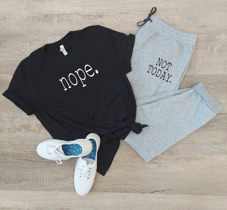 Puede incluir: Una camiseta negra con la palabra "nope" impresa en blanco en la parte delantera, combinada con pantalones de ch&aacute;ndal grises con el texto "NOT TODAY" impreso en negro en la pierna. El conjunto est&aacute; colocado sobre un suelo de madera.