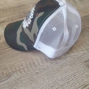May include: A camouflage and white trucker hat with a white daisy on the front.
