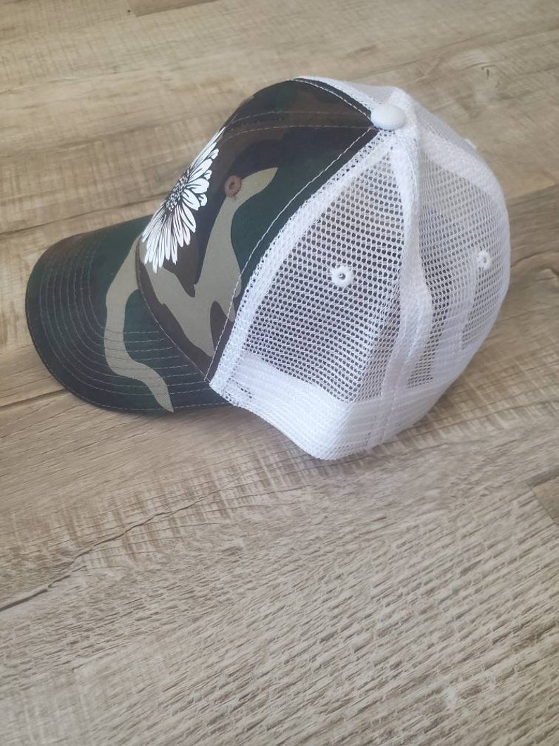 May include: A camouflage and white trucker hat with a white daisy on the front.