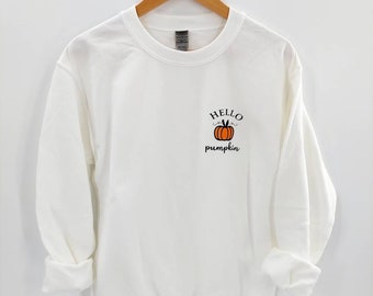 以下が含まれることがあります： 前面に小さなオレンジ色のカボチャのグラフィックと「HELLO pumpkin」の文字が書かれた白いスウェットシャツです。