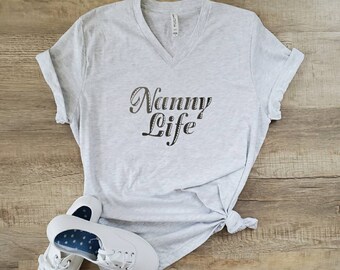 Może przedstawiać: Szary T-shirt z dekoltem w kształcie litery V z nadrukiem "Nanny Life" w kolorze czarnym na przodzie. Koszulka jest zawiązana na dole i leży na drewnianej powierzchni obok pary białych butów sportowych.