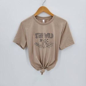 May include: A tan short-sleeved t-shirt with the text "Stay Wild" in black lettering and a floral design.