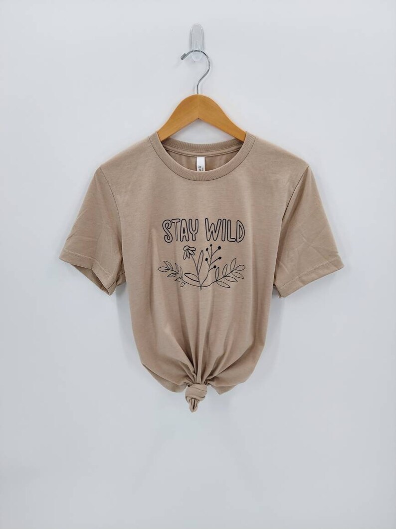 May include: A tan short-sleeved t-shirt with the text "Stay Wild" in black lettering and a floral design.
