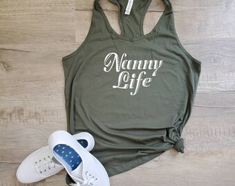 Może przedstawiać: Zielony top na ramiączkach z napisem "Nanny Life" w białym, ozdobnym pismem. Top leży na drewnianej podłodze obok pary białych butów sportowych.