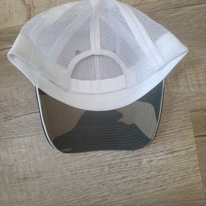 May include: White mesh trucker hat with a camouflage brim. The hat has a white mesh crown and a black brim with a camouflage pattern.