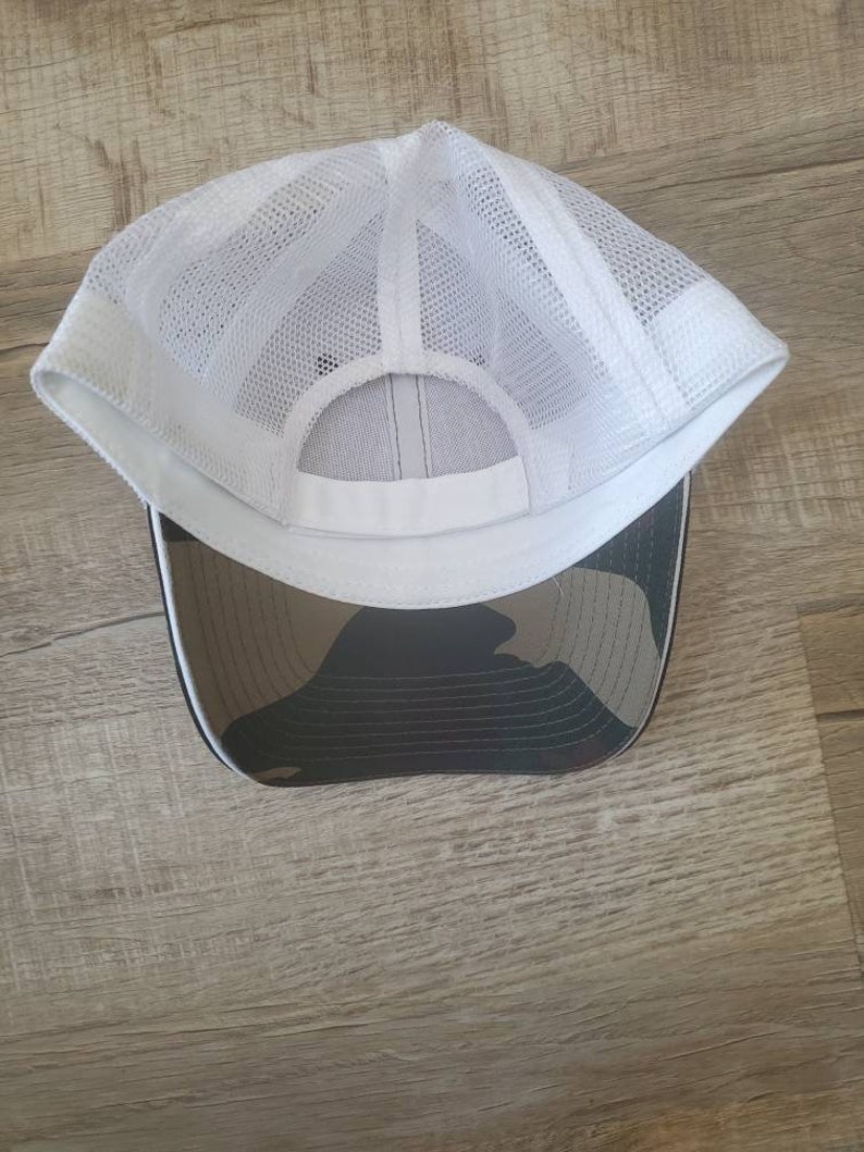 May include: White mesh trucker hat with a camouflage brim. The hat has a white mesh crown and a black brim with a camouflage pattern.