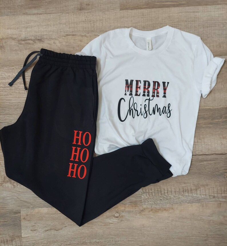 Op de afbeelding: Zwarte joggingbroek met "HO HO HO" in rood op het linkerbeen en een wit T-shirt met "MERRY Christmas" in zwart-rood geruite letters.
