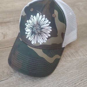 May include: A camouflage trucker hat with a white daisy design. The hat has a white mesh back.