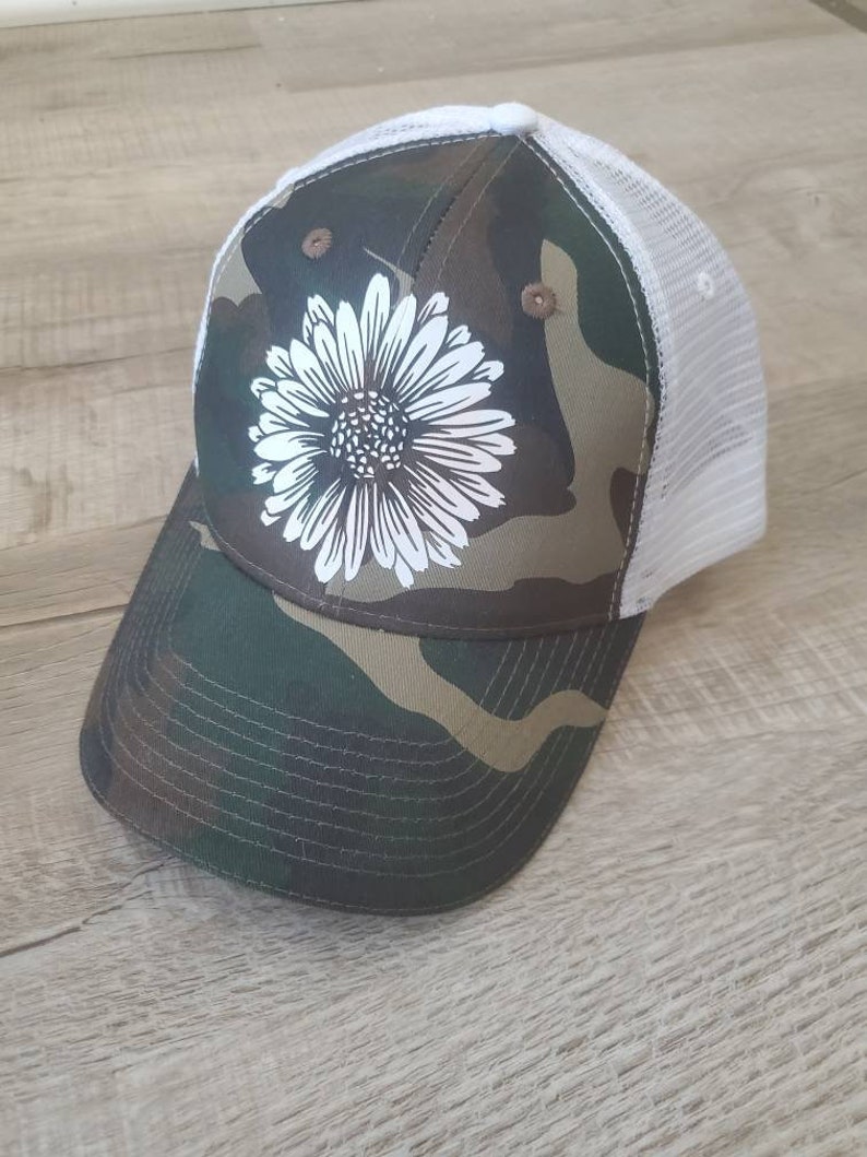 May include: A camouflage trucker hat with a white daisy design. The hat has a white mesh back.