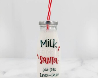 May include: A clear glass milk bottle with a silver lid and a red and white striped straw. The bottle is filled with milk and has the words "Milk for Santa" in red and green, along with names below. A festive Christmas item.