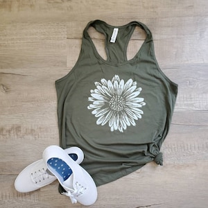 May include: A green tank top with a white daisy graphic. The tank top is made of a soft, comfortable material and has a racerback style. Two white sneakers are also pictured.