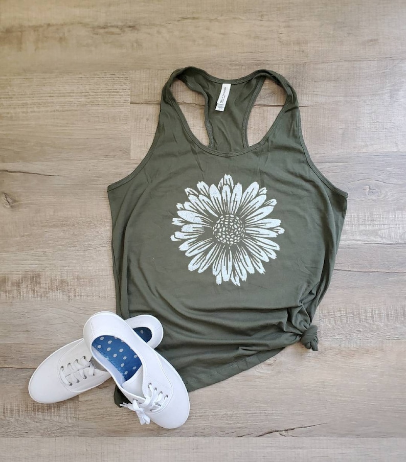 May include: A green tank top with a white daisy graphic. The tank top is made of a soft, comfortable material and has a racerback style. Two white sneakers are also pictured.