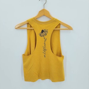 May include: A mustard yellow sleeveless tank top with a black graphic of a wildflower and the text "wildflower" on the back.