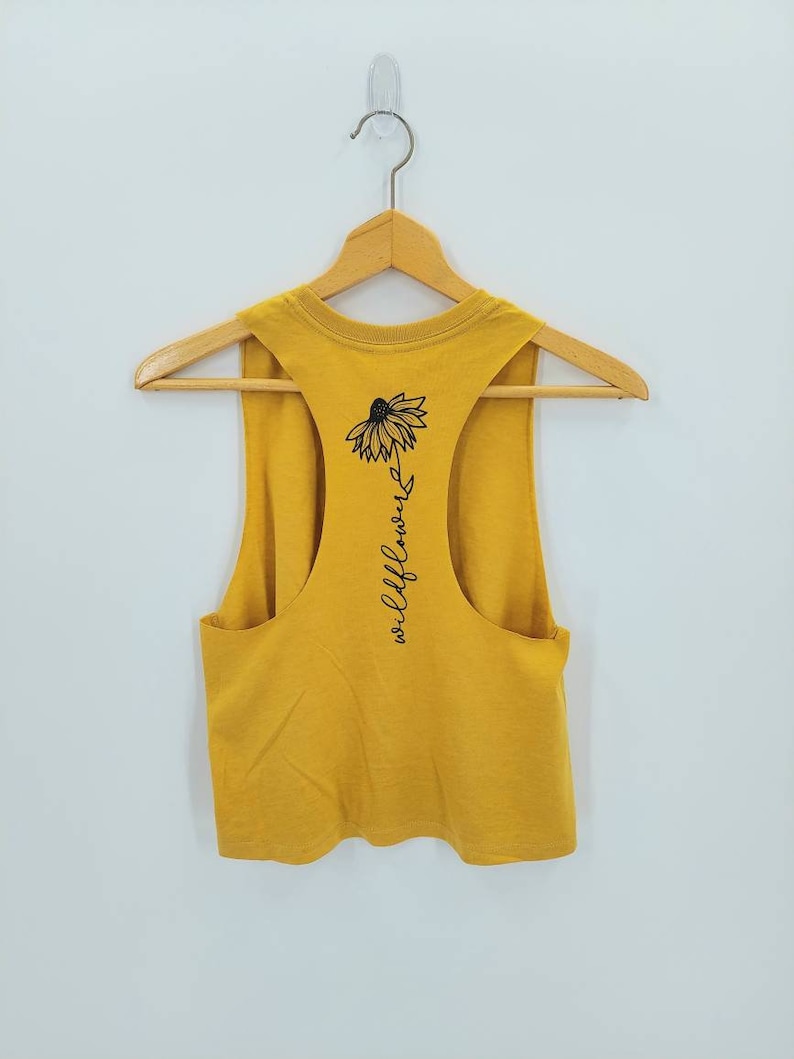 May include: A mustard yellow sleeveless tank top with a black graphic of a wildflower and the text "wildflower" on the back.
