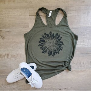 May include: A green tank top with a black sunflower graphic. The tank top has a racerback style. Two white sneakers are on the floor below the tank top.