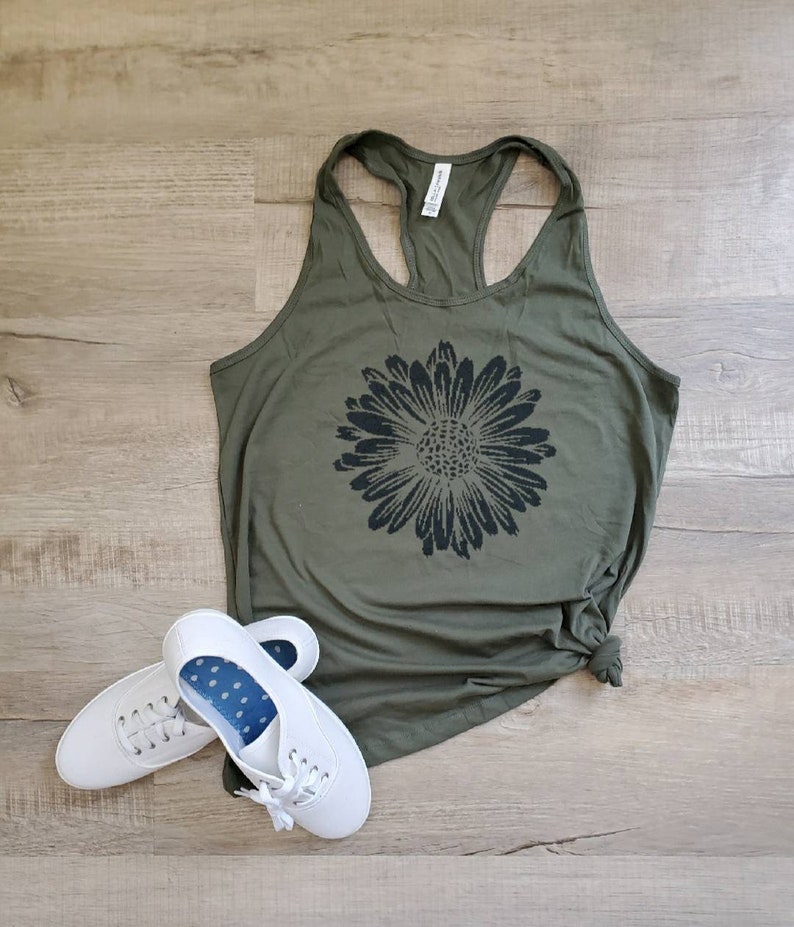 May include: A green tank top with a black sunflower graphic. The tank top has a racerback style. Two white sneakers are on the floor below the tank top.
