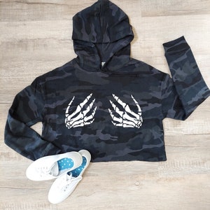 May include: A cropped black and gray camouflage hoodie with a white skeleton hand graphic. The hoodie has a hood and long sleeves. White canvas sneakers with blue polka dot insoles are at the bottom.