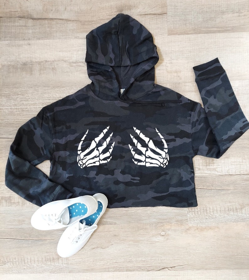 May include: A cropped black and gray camouflage hoodie with a white skeleton hand graphic. The hoodie has a hood and long sleeves. White canvas sneakers with blue polka dot insoles are at the bottom.