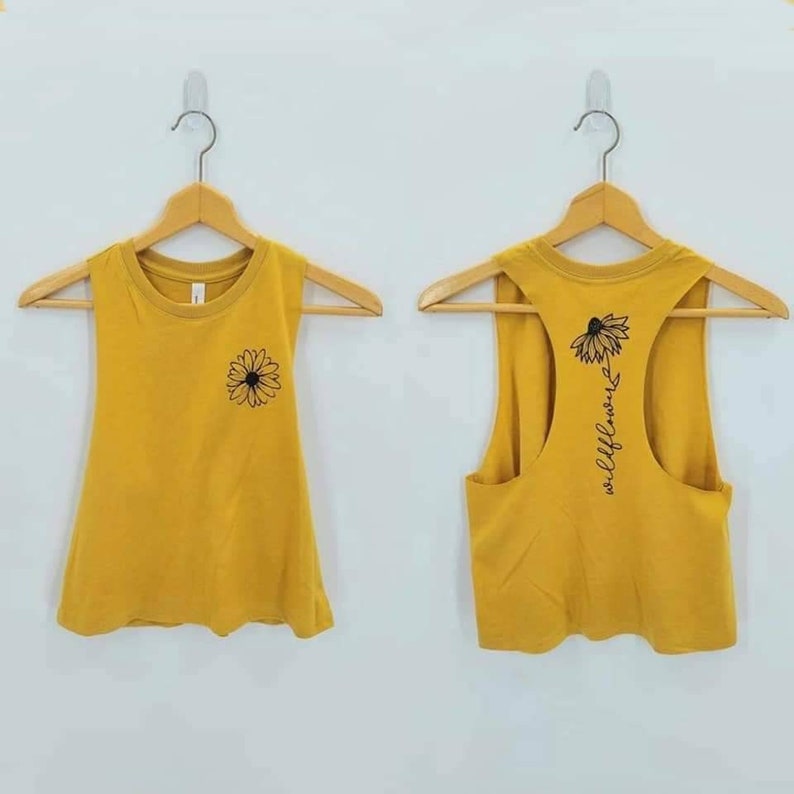 May include: Two mustard yellow tank tops with a black daisy graphic. One tank top has the word "wildflower" written vertically on the back.