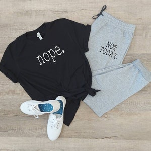 Puede incluir: Una camiseta negra con la palabra "nope" impresa en blanco en la parte delantera, combinada con pantalones de ch&aacute;ndal grises con el texto "NOT TODAY" impreso en negro en la pierna. El conjunto est&aacute; colocado sobre un suelo de madera.