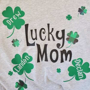Puede incluir: Una camiseta gris con el texto "Lucky Mom" en negro. Hay tr&eacute;boles de cuatro hojas verdes con los nombres "Drew", "Landen" y "Declan" en ellos.