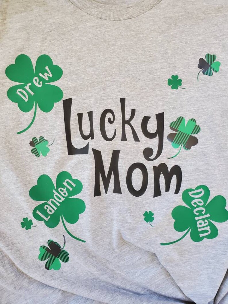 Puede incluir: Una camiseta gris con el texto "Lucky Mom" en negro. Hay tr&eacute;boles de cuatro hojas verdes con los nombres "Drew", "Landen" y "Declan" en ellos.