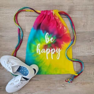 May include: A tie-dye drawstring bag with the words "be happy" in white lettering. The bag is yellow, blue, green, pink, and purple. Two white canvas shoes are on the wooden surface next to the bag.
