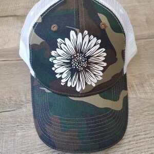 May include: A camouflage trucker hat with a white daisy design. The hat has a white mesh back.