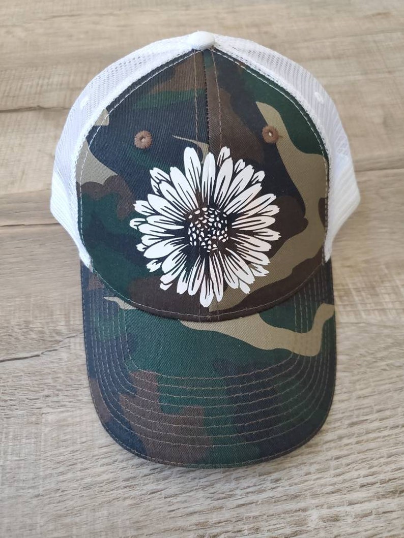 May include: A camouflage trucker hat with a white daisy design. The hat has a white mesh back.