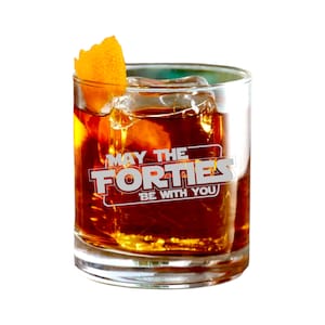 May include: A tumbler glass containing a dark amber cocktail, ice cubes, and an orange peel garnish. The glass features the text "MAY THE FORTIES BE WITH YOU" in a white, stylized font.