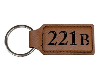 Pode incluir: Chaveiro de couro marrom com um chaveiro de prata e o texto "221B" gravado na frente.