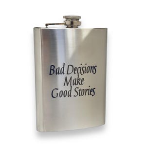 Pode incluir: Uma garrafa de bolso de aço inoxidável prateada com o texto "Bad Decisions Make Good Stories" gravado na frente.