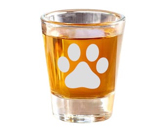 Puede incluir: Un vaso de chupito de vidrio transparente con un diseño de huella de pata blanca lleno de líquido ámbar.