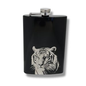May include: Black stainless steel hip flask with a silver cap and hinge. The front displays a black and white tiger face. The flask is rectangular with a curved top. Measures approximately 12 cm in height.