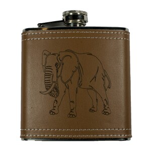 May include: A brown leather-covered flask with an engraved image of an elephant. The flask has a silver top and a stitched edge.