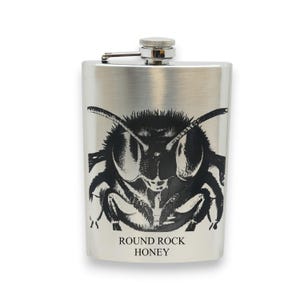 May include: A stainless steel flask with a detailed black and white illustration of a bee's face. The words "ROUND ROCK HONEY" are printed below the image. The flask has a silver cap and a hinged closure.