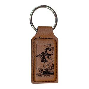 May include: Brown leather keychain with a silver key ring. The keychain features a rectangular leather tag with an engraved image of The Fool tarot card. The text "THE FOOL." is printed below the image.
