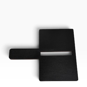 Puede incluir: Un soporte de exhibición plano, negro y rectangular de madera que presenta una ranura horizontal cortada en el centro del panel principal y un brazo rectangular que sobresale del lado izquierdo, diseñado para sostener objetos en posición vertical.
