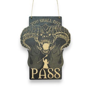 May include: Wooden sign with a fantasy theme. The sign features a detailed illustration of a demonic creature with horns and flames, along with the text "YOU SHALL NOT PASS" and "PASS". The sign is suspended by a natural twine.