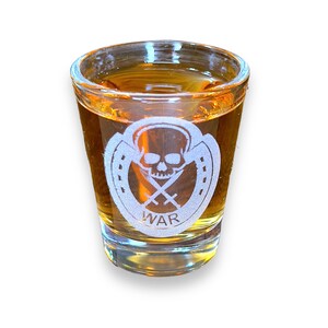 Peut inclure: Un verre à shot en verre transparent rempli d'un liquide doré. Le verre est gravé d'un motif blanc représentant un crâne avec des épées croisées et le mot "WAR" en dessous.