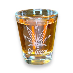 Puede incluir: Un vaso de chupito de vidrio transparente con un diseño de hoja de marihuana verde y el texto "Go Green" grabado en el lateral. El vaso está lleno de un líquido dorado.