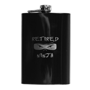 May include: A black stainless steel flask with a hinged cap. The flask has the words "RETIRED NINJA" and a ninja mask design etched on the front. The flask is rectangular and has a shiny finish.