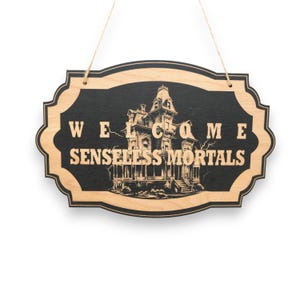 May include: Wooden Halloween sign with a black background and a light wood border. The sign reads "WELCOME SENSELESS MORTALS" with a haunted house illustration. The sign is hanging by a natural twine.