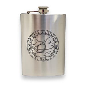 May include: A silver stainless steel hip flask with a black and white engraved design that reads "Houston, we have a drinking problem" with a space-themed graphic. The flask is made in the U.S.A.