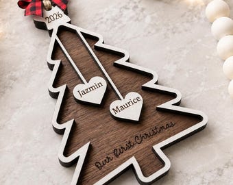 May include: A wooden Christmas tree ornament featuring a dark brown center and white border. It includes a red and black buffalo plaid bow, the year 2026, two white heart-shaped tags engraved with Jazmin and Maurice, and the text Our first Christmas.