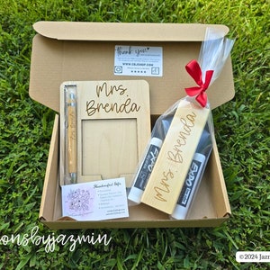 May include: A gift box containing a wooden picture frame, pen, and bookmark, all engraved with "Mrs. Brenda." The box also includes two Expo dry-erase markers. The items are arranged on a bed of grass.