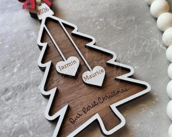 May include: Wooden Christmas tree ornament with a white outline and a dark wood grain center. The ornament has two heart-shaped cutouts with names, the year 2026, and the phrase "Our first Christmas." A red and black plaid bow tops the ornament.