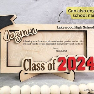 May include: Wooden graduation photo frame with the name "Dazmin" and "Class of 2024" in red. The frame includes the text "Lakewood High School" and a graduation cap. The frame has a stand and includes a message from Mom & Dad.